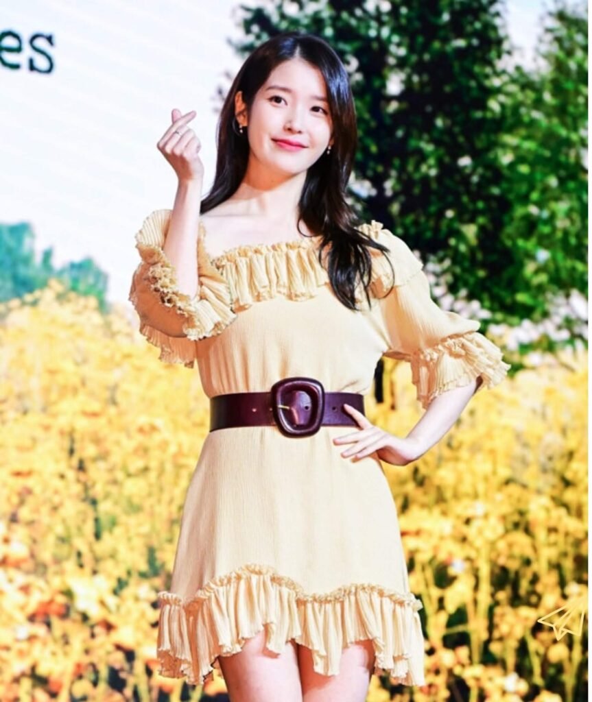 IU wearing a yellow one-piece dress smiling while making a heart with her hands